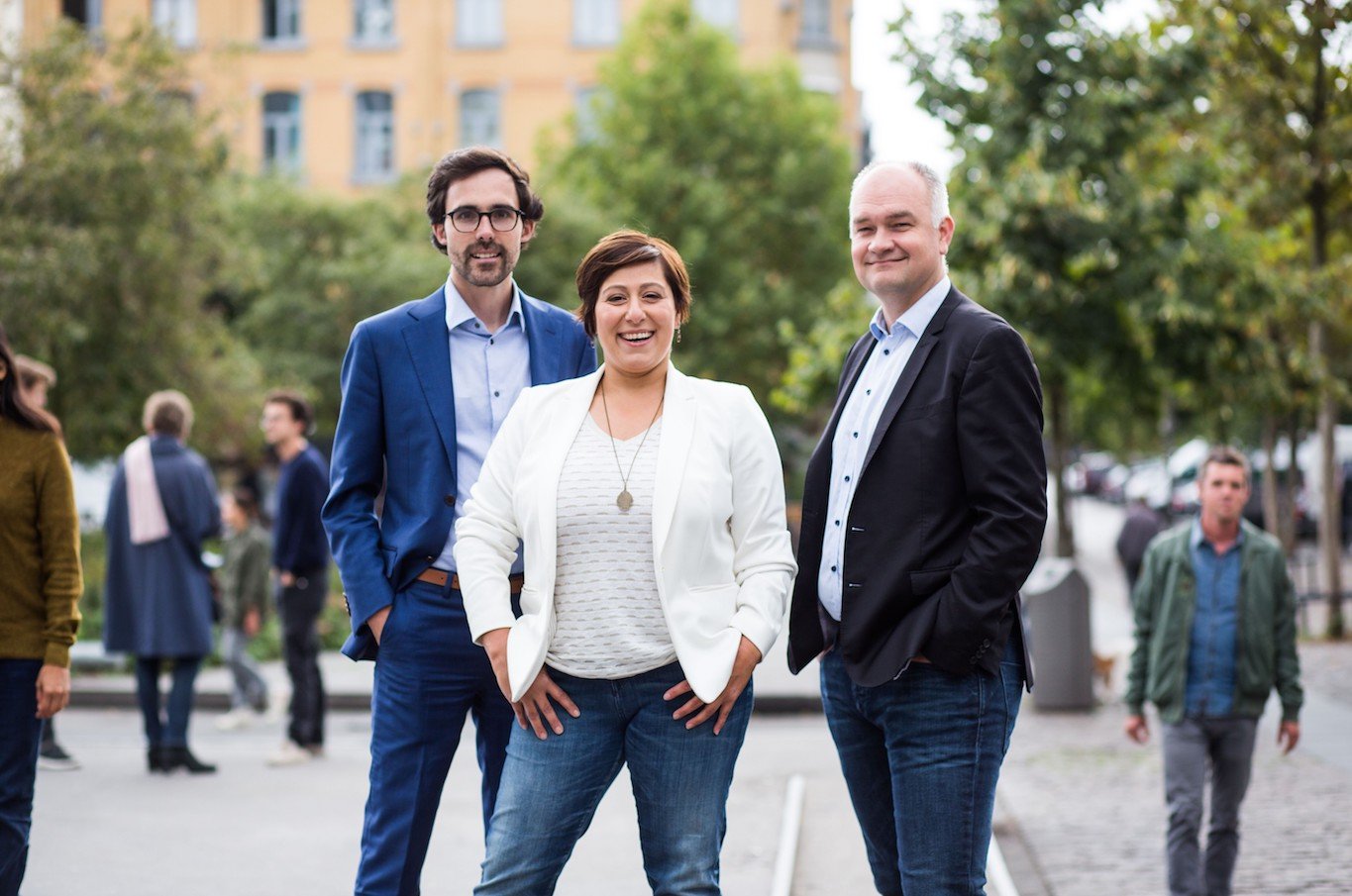 A group of three professional colleagues—two men in blazers and a woman in a white jacket—posing together in a European city square.