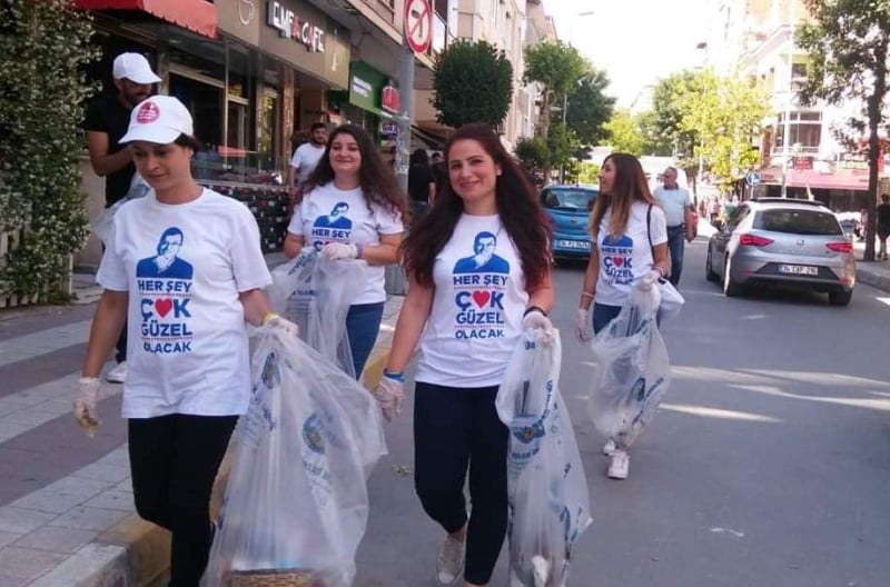 A group of women wearing matching shirts and gloves walk down a city street carrying plastic bags filled with collected litter during a cleanup event.