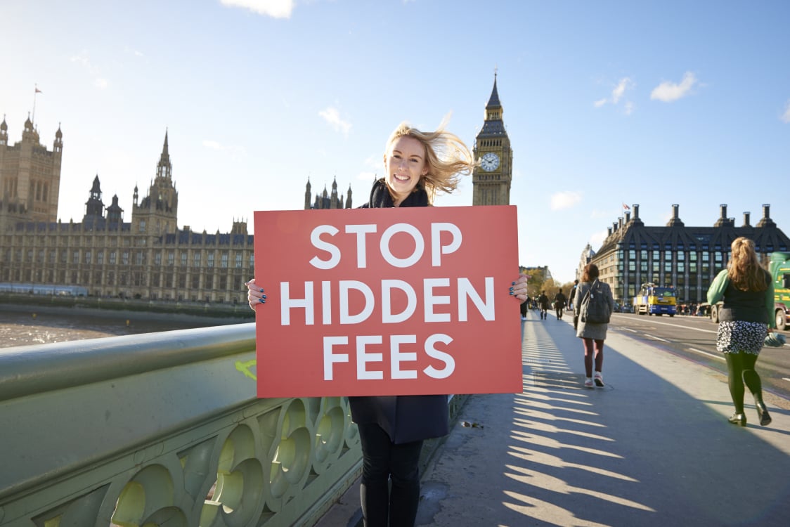 A woman holds a red sign reading “Stop hidden fees” on Westminster Bridge in London during a consumer rights campaign.