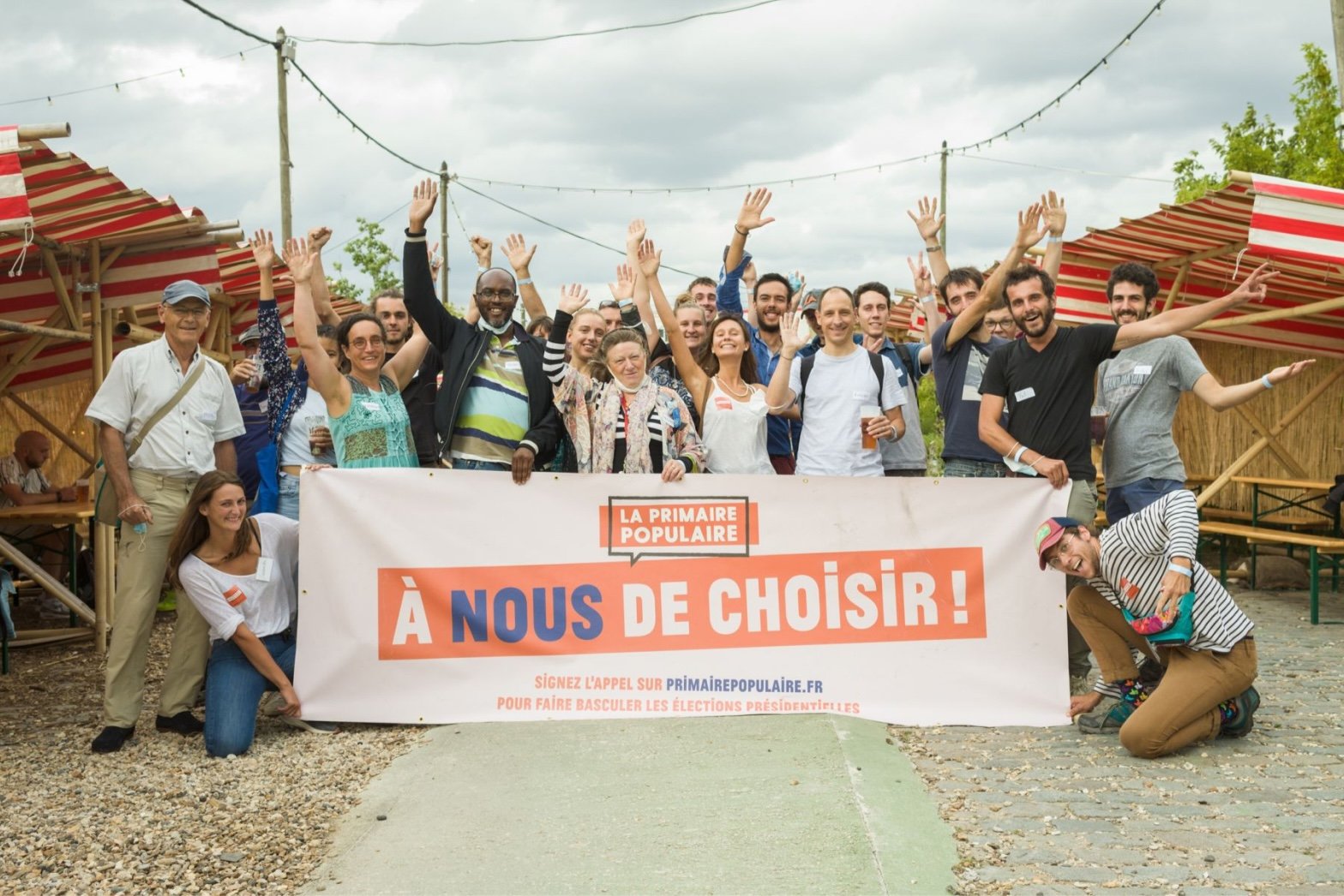 A smiling group of people pose outdoors behind a banner that reads “À nous de choisir!” at a community event organized by La Primaire Populaire.