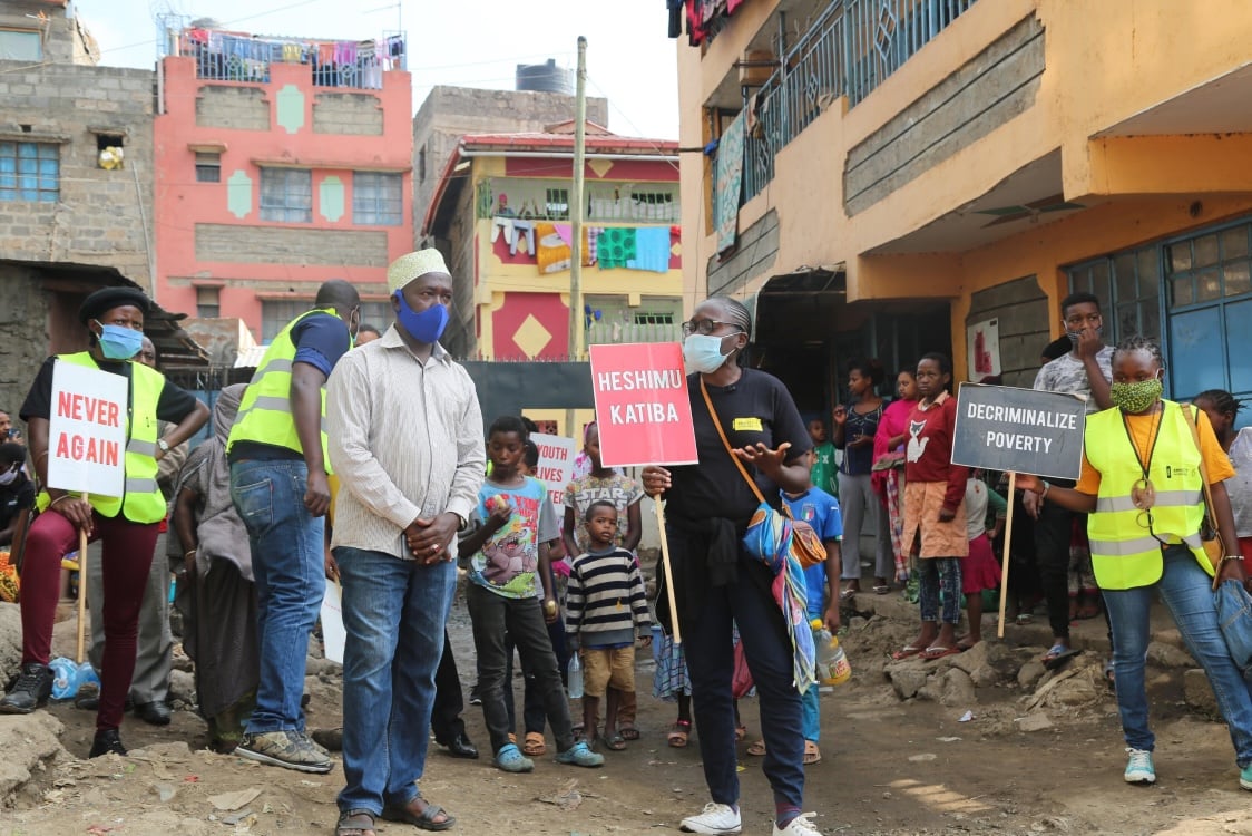 People wearing reflective vests and masks hold protest signs in a residential area. Signs read “Never again,” “Heshimu Katiba,” and “Decriminalize poverty.”